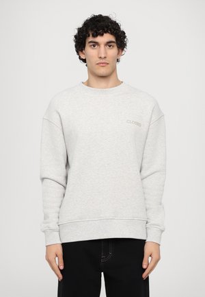 Sweatshirt gris clair à manches longues, col rond, avec des poignets et un ourlet côtelés ; présente une broderie subtile "CLOSED" sur la poitrine. Jean noir.