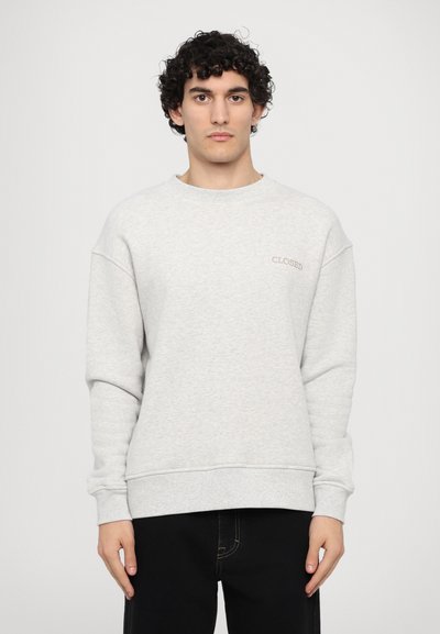 Hellgrauer Pullover mit langen Ärmeln, Rundhalsausschnitt, gerippten Bündchen und Saum; mit subtiler "CLOSED"-Stickerei auf der Brust. Schwarze Jeans.