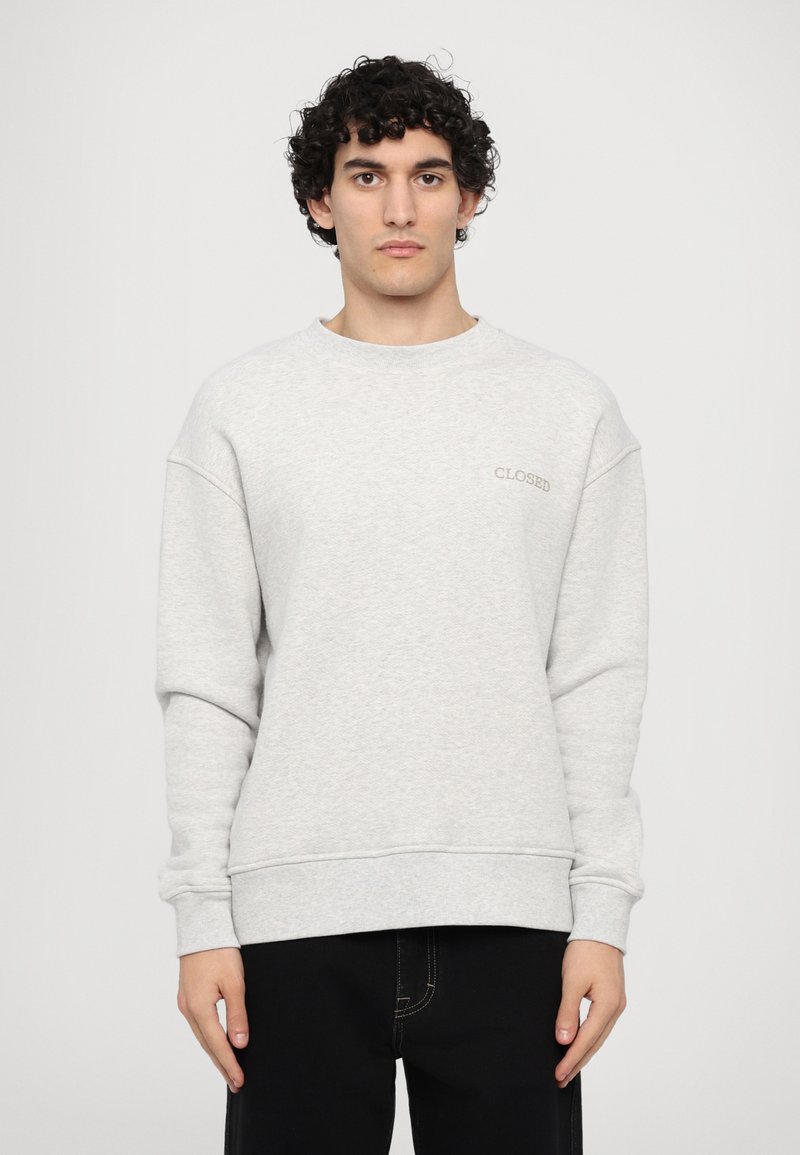 Light gray sweatshirt with long sleeves, round neck, ribbed cuffs and hem; features subtle "CLOSED" embroidery on the chest. Black jeans.