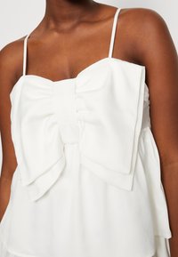 Haut blanc avec un grand nœud au centre, fines bretelles et coupe ample. Tissu lisse avec un éclat subtil et détails de design en couches.