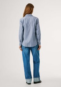 Camicia a maniche lunghe blu e bianca a righe con orlo curvo, polsini abbottonati, in morbido cotone, indossata con jeans azzurri a vestibilità morbida.