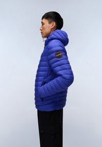 Giacca imbottita blu con cappuccio, caratterizzata da sezioni trapuntate orizzontali, tasche laterali e un patch circolare con logo sulla spalla sinistra.