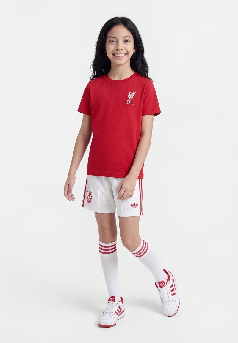 Χαμογελαστή κοπέλα με κόκκινη μπλούζα Liverpool FC, άσπρες βερμούδες με το λογότυπο της ομάδας, άσπρες κάλτσες μέχρι το γόνατο με κόκκινες ρίγες και άσπρα αθλητικά παπούτσια Adidas.