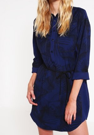 Woman wearing a navy blue long-sleeve shirt dress with a black belt tied at the waist and subtle dark leaf patterns.