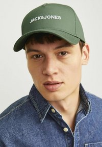 Grøn baseballcap med "JACK & JONES" logo i hvid. Glat stof, struktureret form, buet skygge. Båret af en person i en denimskjorte.