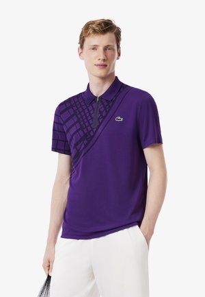 Lila Polo-Shirt mit kurzen Ärmeln, ausgestattet mit einem diagonalen schwarzen Karomuster und einem Reißverschlusskragen. Lacoste-Logo in Grün auf der Brust.