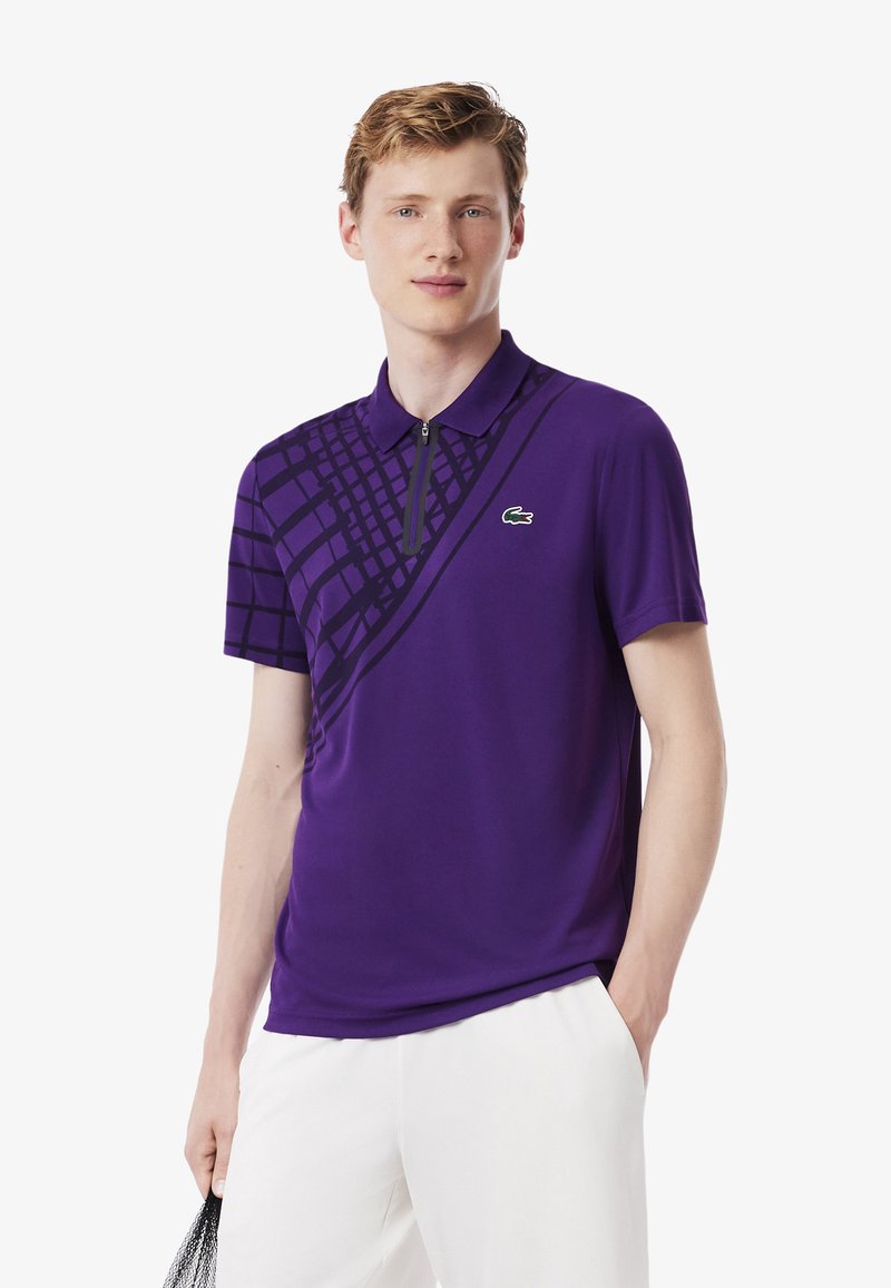 Paarse polo shirt met korte mouwen, voorzien van een diagonaal zwart ruitpatroon en een ritssluiting bij de kraag. Lacoste-logo in het groen op de borst.
