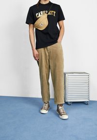 Černé tričko s zlatým nápisem "Carhartt", světlé manšestrové kalhoty a hnědé vysoké tenisky. Černá křížová taška a stříbrná úložná krabice v pozadí.