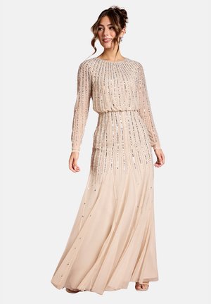 Donna che indossa un lungo vestito beige con decorazioni verticali di paillettes e tessuto trasparente, in piedi con un leggero sorriso e i capelli raccolti in uno chignon morbido.