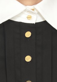 Indumento nero con pieghe verticali, dotato di un colletto bianco. Chiuso con bottoni dorati, che mostra una texture di tessuto liscia.