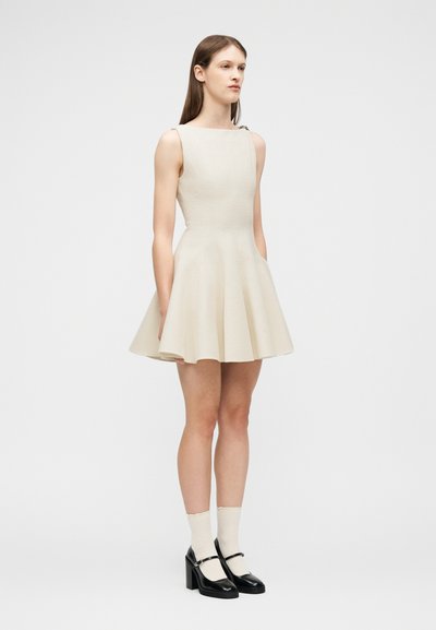 Γυναίκα που στέκεται φορώντας ένα αμάνικο μπεζ φαρδύ φόρεμα, λευκές κάλτσες και μαύρα παπούτσια πλατφόρμας Mary Jane μπροστά σε ένα απλό λευκό φόντο.