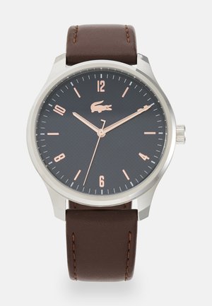 Round watch with a black textured dial, silver case, and rose gold hour markers. Brown leather strap with contrasting stitching.