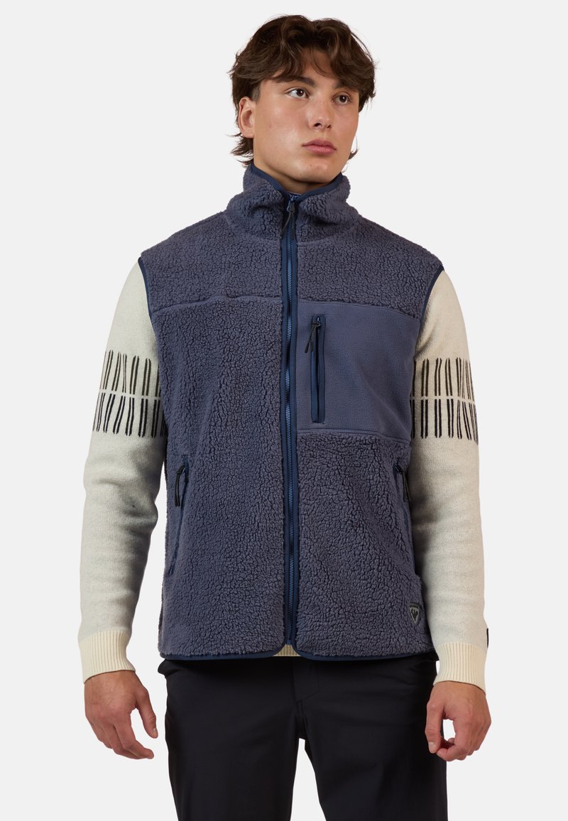 Fleeceväst med hög krage, blå och beige färgkontrast, ficka med dragkedja på bröstet, texturerad kropp och randiga ärmdetaljer.