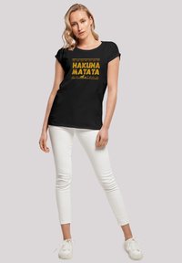 Svart t-skjorte med gult "HAKUNA MATATA"-tekst og geometriske mønstre. Kombinert med hvite tettsittende bukser og hvite sneakers.