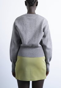 Personne aux cheveux tressés portant des boucles d'oreilles anneaux argentées, un pull en maille gris et une mini-jupe en cuir vert clair, vue de dos.