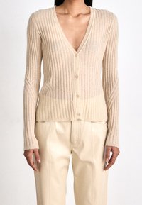 Beige pletený kardigan s hlubokým V-výstřihem, zapínáním na pět knoflíků a roztrhanými okraji. Nosený přes lesklé krémové kalhoty.