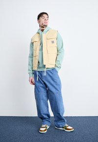 Beige vesta s několika předními kapsami přes světle zelenou mikinu, spárovaná s volnými modrými džíny a vícebarevnými teniskami.