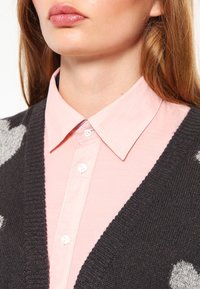 Šedý pletený kardigan s bílými puntíky, nošený přes světlou růžovou košilí s límečkem. Kardigan má V-výstřih a ribované okraje.