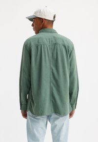 Camisa de manga larga verde hecha de un material suave, con un cuello abotonado, costura en el yugo trasero y dobladillo curved. Combinada con unos vaqueros azules claros.