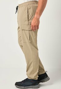 Pantaloni cargo beige realizzati in tessuto leggero, con vita elastica, tasche laterali e vestibilità comoda. Sneakers visibili sotto.