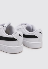 Sneakers bianchi in materiale sintetico, con una striscia nera, tacco arrotondato e un logo sul lato. Suola in gomma per migliorare la presa.