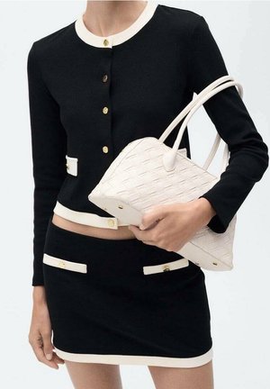 Femme portant un cardigan boutonné noir et blanc avec un ensemble jupe, tenant un sac à main en cuir tressé blanc avec des doubles poignées.