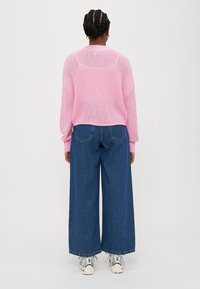 Rozā adīta džemperis ar vaļēju austību, saskaņots ar platām zilu džinsu biksēm. Apģērbu pabeidz baltas kedas, kas sniedz neformālu stilu.