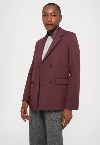 Sieviete, tērpusies divrindu burgundijas krāsas žaketē virs gaismas džemperi un pelēkiem teksturētiem biksēm, stāvot pretī vienkāršai baltai fonam.