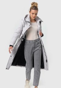 Lång, ljusgrå pufferjacka med huva, svart insida och dragkedjestängning, bärs över en beige topp och grå jeans, matchad med vita sneakers.