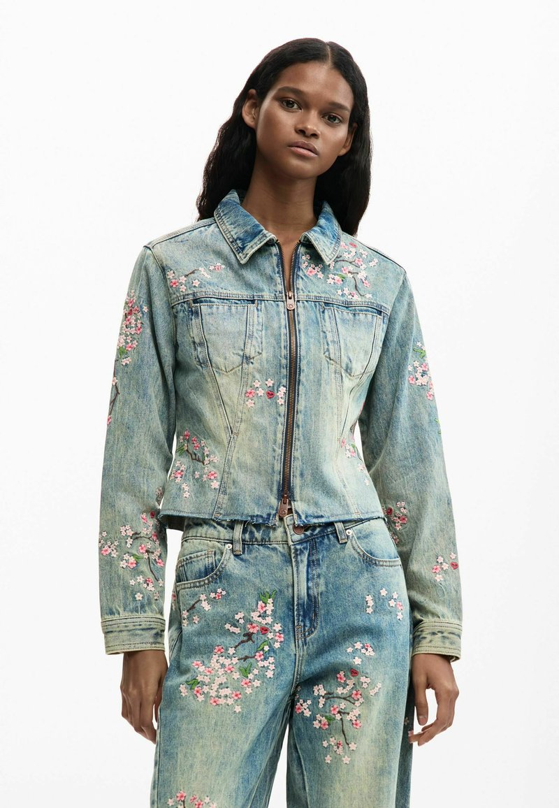 Femme portant une veste en jean bleu clair et un jean avec une broderie florale rose et blanche, debout devant un fond blanc uni.