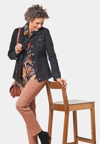 Dunkelblaue Jeansjacke mit silbernen Knöpfen, zwei vorderen Taschen, kombiniert mit einer mehrfarbigen gemusterten Bluse und beigen Hosen. Holzstuhl im Hintergrund.