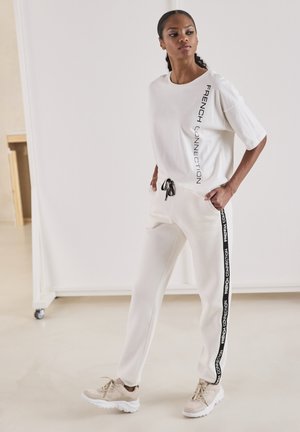 Weißes Kurzarm-T-Shirt mit dem Aufdruck "FRENCH CONNECTION", kombiniert mit cremefarbenen Jogginghosen mit seitlichen Streifen und beigen Sneakers.