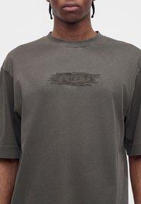 Άτομο που φοράει σκούρο γκρι oversized μπλουζάκι με υφασματικό αφηρημένο λογότυπο στο κέντρο του στήθους και κοντά μανίκια.