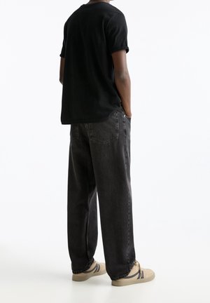 Personne portant un t-shirt noir, un jean noir ample et des baskets beiges avec des rayures bleu marine, debout les mains dans les poches, de dos.