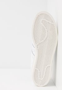 Zapatilla blanca con suela de goma texturizada que presenta patrones en zigzag. Rayas laterales en gris, parte superior de cuero suave y diseño minimalista.