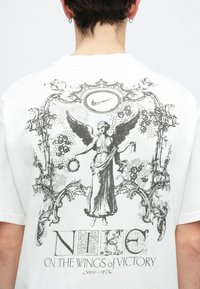 Biely bavlnený tričko s čiernym grafickým tlačom anjela držiaceho vavrín, orámované dekoratívnymi prvkami a logom Nike.