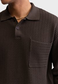 Polo marrón con textura y un patrón ondulado, con cuello, dos botones morados y un bolsillo parche en el pecho izquierdo.