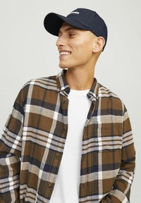 Brown and beige plaid shirt with white and navy accents, buttoned at the collar, paired with a navy cap featuring embroidered text.