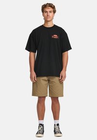 Svart bomullströja med en liten röd Billabong-logotyp; kombinerad med khakifärgade shorts och svarta högskor, med vita strumpor.