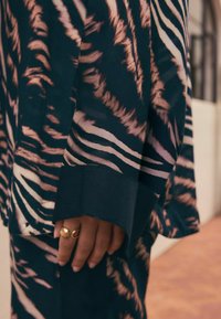 Tissu noir et beige à motif rayé tigre. Présente une manche ample avec un poignet uni de couleur sombre et une main portant des bagues en or.