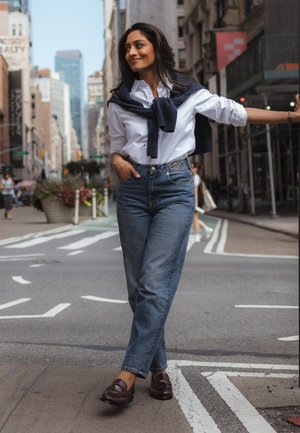 Une femme en chemise blanche, en jean et en mocassins se tient en souriant sur un passage piéton en ville, un pull bleu marine posé sur les épaules.