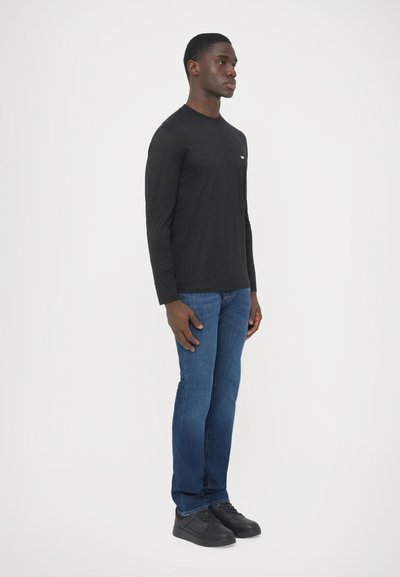 Fekete, hosszú ujjú póló kerek nyakkivágással, kék farmernadrággal és fekete sportcipővel párosítva. A pólón a bal mellkason egy kis logó található.