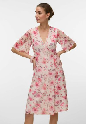 Femme portant une robe portefeuille fleurie rose et beige longueur genou avec manches aux coudes, debout les mains sur les hanches devant un fond uni.
