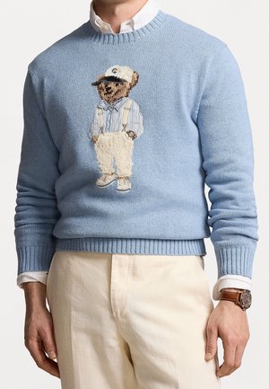 Mann trägt hellblauen Pullover mit besticktem Teddybär mit Hosenträgern und Mütze, kombiniert mit beigen Hosen und einer Armbanduhr.
