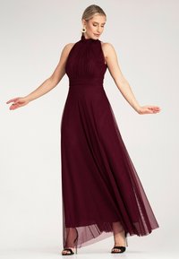 Burgunderfarbenes Maxikleid mit Halterneck, plissiertem Oberteil, fließendem Rock und transparenter Überlage. Wird mit schwarzen Sandalen mit Absatz kombiniert. Glatte Textur.