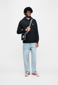 Μαύρο φούτερ με κουκούλα και λογότυπο, ανοιχτό μπλε τζιν παντελόνι φαρδιάς γραμμής και κόκκινα αθλητικά παπούτσια. Μαύρη τσάντα χιαστί με λευκό λογότυπο αστεριού και ριγωτό λουρί.