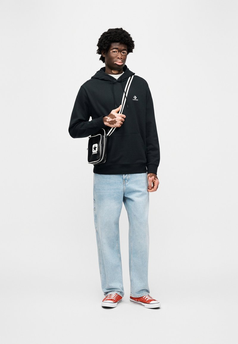 Hoodie noir avec logo, jean large à jambes larges bleu clair, et baskets rouges. Sac bandoulière noir avec un logo étoile blanche et une bandoulière rayée.