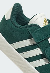 Sneaker in camoscio verde con dettagli in pelle bianca e tre strisce bianche. Caratterizzato da una suola in gomma testurizzata e un cinturino con chiusura a velcro.