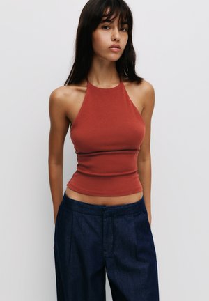 Halter top u rust boji s prilagođenim dizajnom, napravljen od glatkog materijala. Uparen s visokim tamnoplavo širokim hlačama.