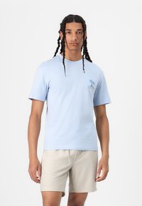 Light blue cotton t-shirt with short sleeves, featuring a small palm tree graphic and brand name on the chest. Paired with beige textured shorts.